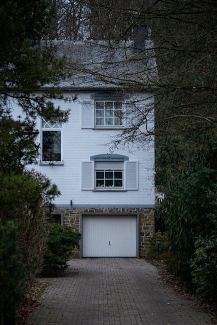 A white, three-storey detached house with a stone base and a garage on the ground level, situated at the end of a paved driveway surrounded by trees and shrubbery, during daylight hours. The house fea